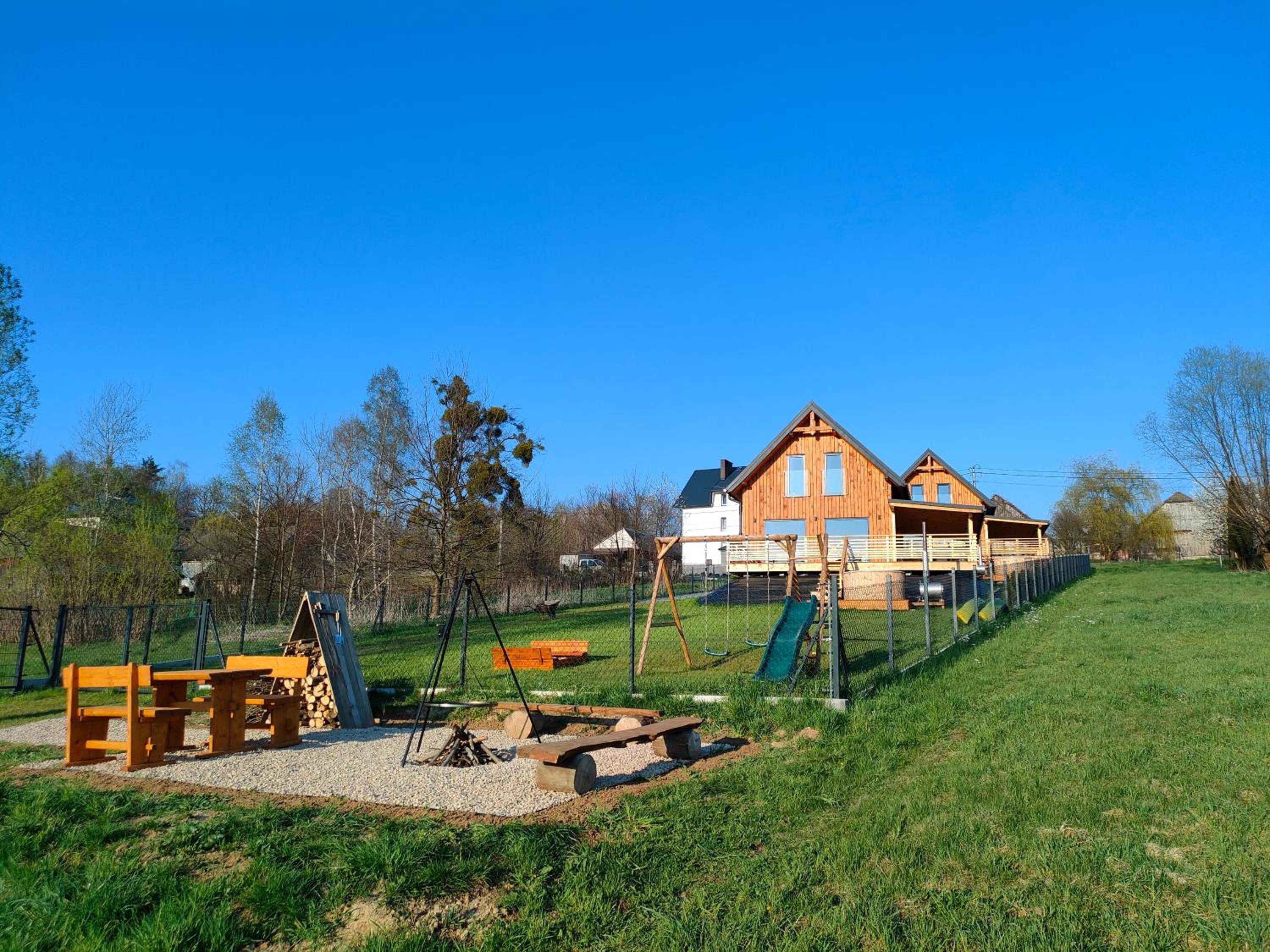Drewniany domek na zielonej łące, przed nim plac zabaw i miejsce na ognisko z ławkami